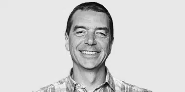 A man with short dark hair, wearing a plaid shirt, smiles at the camera against a plain light gray background.